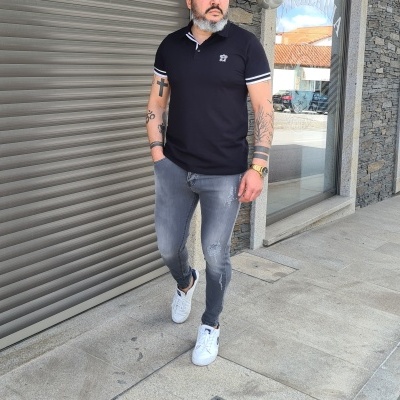 Homem com polo preta, jeans cinza e ténis brancos a caminhar numa calçada urbana