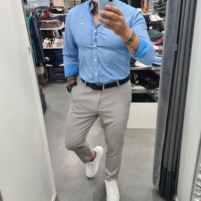 Homem com camisa azul clara de padrão, calças bege, cinto preto e sapatos brancos em loja de roupas