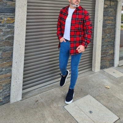 Homem com camisa de flanela xadrez vermelha e preta, t-shirt branca, jeans azul e ténis pretos em frente a porta metálica