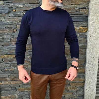 Homem com camisola azul escura e calças castanhas em frente a parede de pedras