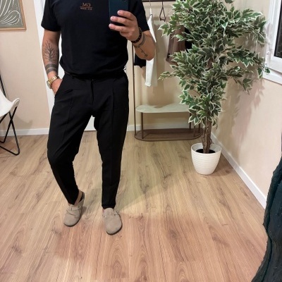 Homem a vestir camiseta preta com texto dourado GM EST. 2000 e calças pretas, com planta verde e branca e cabide de roupa ao fundo