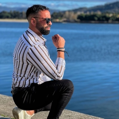 homem agachado junto a rio com camisa às riscas, calças pretas e ténis brancos