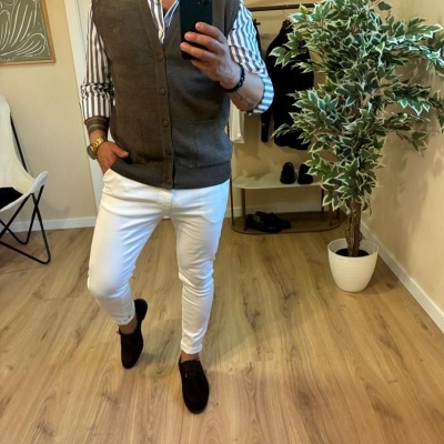 Homem de camisa riscada branca e azul, colete cinzento, calças brancas e sapatos pretos em ambiente interior