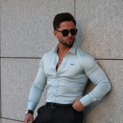 Homem com camisa azul clara e óculos de sol encostado numa parede de pedra cinzenta