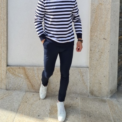 Homem com camiseta de riscas brancas e azuis, calças azul-escuras e sapatos brancos em fundo de parede de pedra