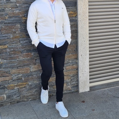 Homem com casaco branco, calças pretas e ténis brancos encostado a parede de pedra