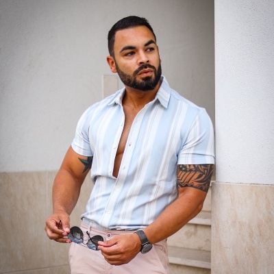 Homem com camisa listrada branca e azul clara, calças bege, tatuagens e a segurar óculos escuros.