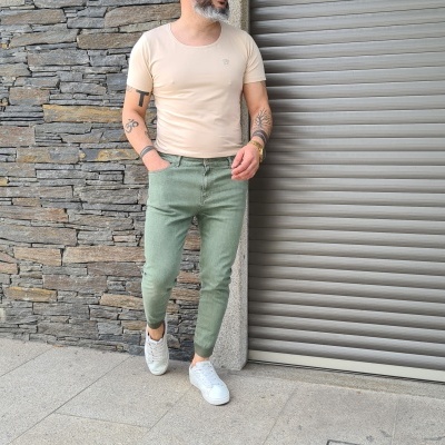 Homem vestido com camiseta bege clara, calças verdes e sapatos brancos junto a parede de pedra e porta metálica