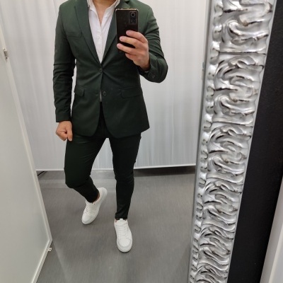 Homem com fato verde escuro, camisa branca e ténis brancos a tirar selfie num espelho com moldura prateada