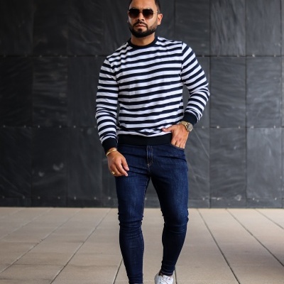 Homem com camisola de riscas pretas e brancas, jeans azul escuro e sapatilhas brancas em piso claro perante parede preta