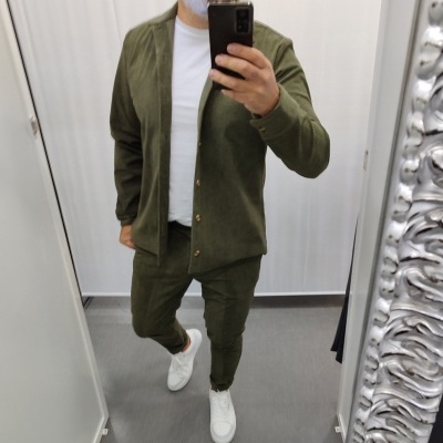 Homem a vestir conjunto verde com camisa de botões e calças, sapatos brancos, tirado em espelho num provador