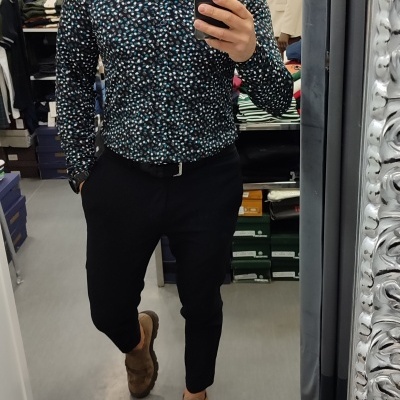 Homem com camisa estampada e calças pretas em loja de roupas fotografando no espelho.