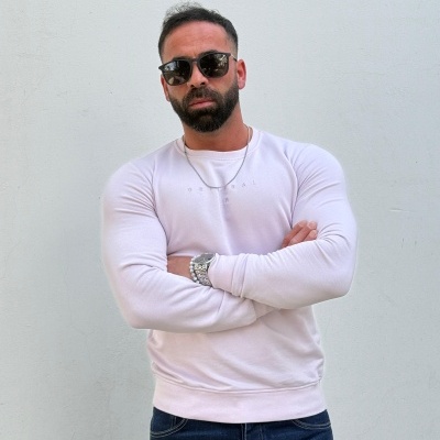 Homem com camisa branca de manga comprida e óculos escuros posando contra parede branca