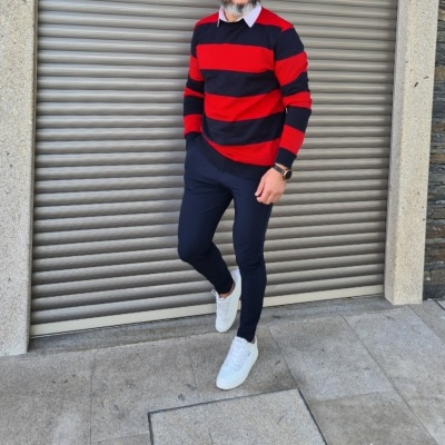 Homem com camisola de riscas vermelhas e pretas, calças azuis e sapatilhas brancas junto a porta metálica