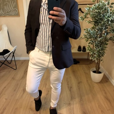 Homem com casaco preto, camisa às riscas azuis e calças brancas a tirar selfie