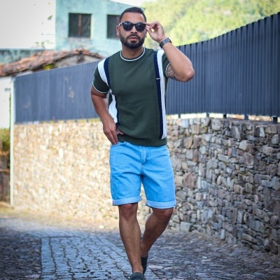 Homem com camisola verde, calções de ganga e ténis a caminhar numa rua de calçada