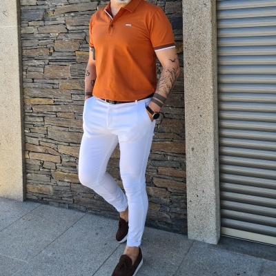 Homem com polo laranja e calças brancas junto a parede de pedra