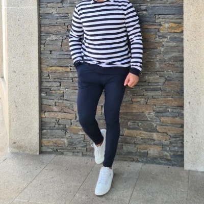 Homem com camisola riscada preta e branca, calças pretas e ténis brancos em frente a parede de pedra