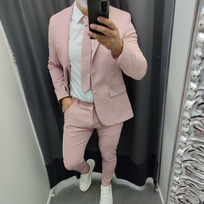 Homem com fato rosa claro, camisa branca, gravata rosa e ténis brancos a tirar selfie em espelho com cortinado cinzento