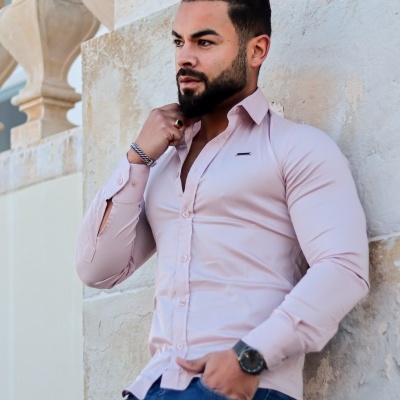 Homem com camisa rosa claro e calças azul escuro encostado a parede de pedra clara