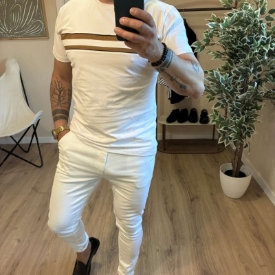 Homem com t-shirt branca e riscas castanhas, calças brancas e sapatos pretos num ambiente interior