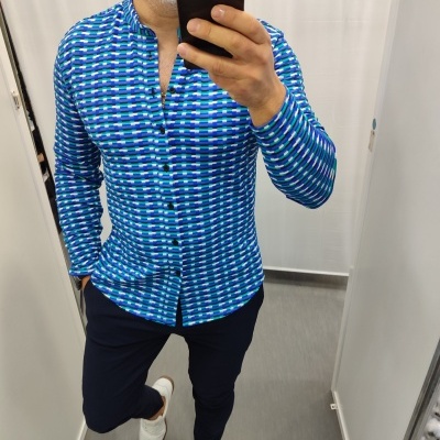 Homem a usar camisa azul com padrão geométrico, calças escuras e ténis brancos, numa cabine de prova.