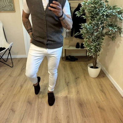 Homem com colete castanho e calças brancas em quarto com planta e cabide de roupa