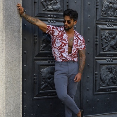 Homem com camisa floral vermelha e branca e calças cinzentas junto a porta preta decorada