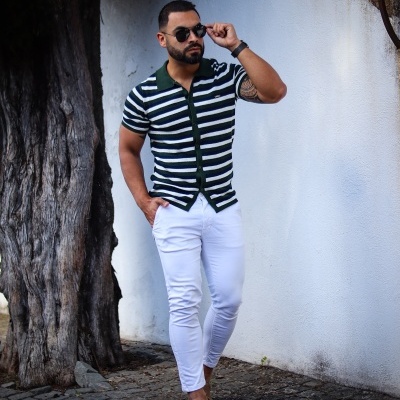 Homem de camisa polo listrada, calças brancas e óculos escuros junto a uma árvore e muro branco.