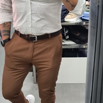 Homem com camisa branca estampada, calças castanhas e sapatos brancos a tirar selfie num espelho numa loja de roupa