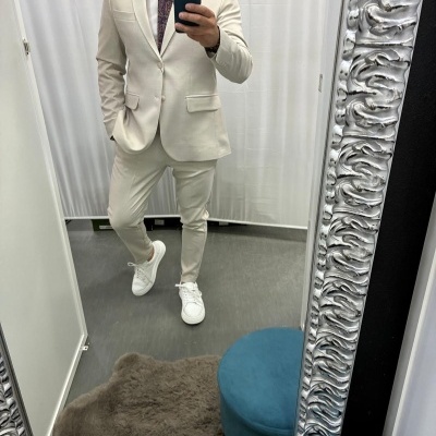 Homem com fato bege claro, gravata bordô e sapatilhas brancas a tirar selfie num espelho com moldura prateada detalhada.