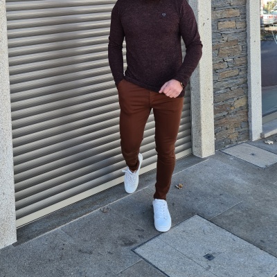 Homem com camisola castanha escura, calças castanhas e ténis brancos junto a porta metálica e parede de pedra