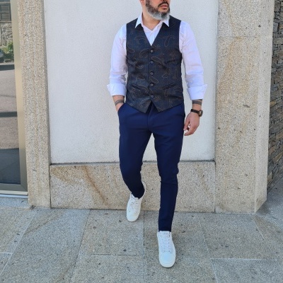 Homem vestido com colete azul escuro com padrão e calças azuis escuras em fundo de parede branca e pedra