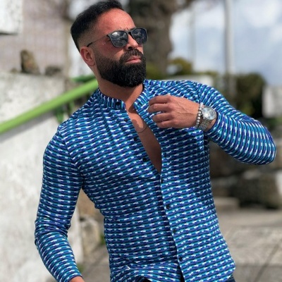 Homem com camisa azul padrão geométrico, relógio prateado, pulseira e óculos de sol em ambiente exterior