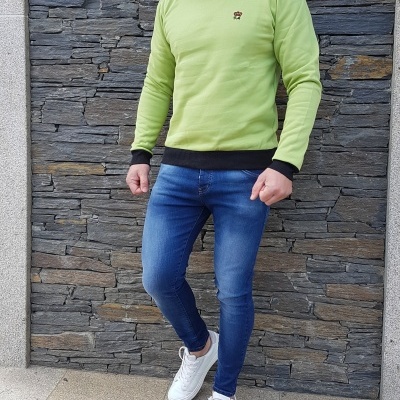 Homem com sweatshirt verde claro e calças de ganga azuis junto a parede de pedra cinzenta