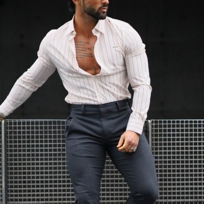 Homem com camisa branca riscada, calças cinzentas e botas pretas encostado numa grade metálica