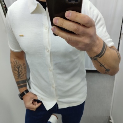 Camisola branca de manga curta com botões e logótipo dourado, usada por homem com tatuagens, num provador