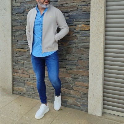 Homem com camisa azul clara, casaco bege, jeans azuis e sapatilhas brancas encostado a parede de pedra cinzenta