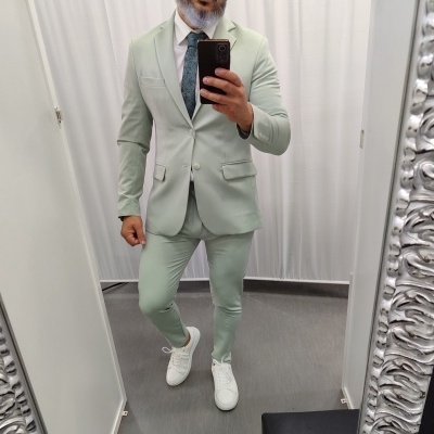 Fato verde-claro de homem com camisa azul, gravata e ténis brancos num provador com espelho decorado.