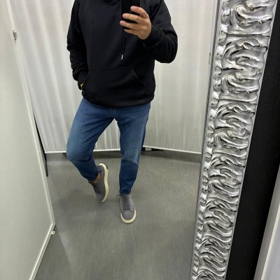 Homem a tentar uma roupa composta por sweatshirt preta com capuz, jeans azuis e sapatilhas cinzentas num provador.