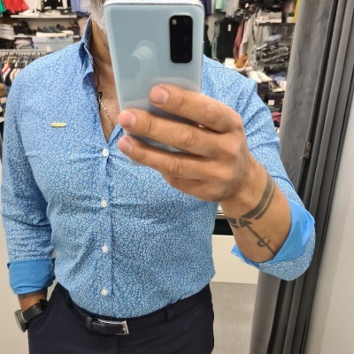 Homem a tirar uma selfie usando camisa azul clara com padrão floral branco e calças pretas na loja de roupas.