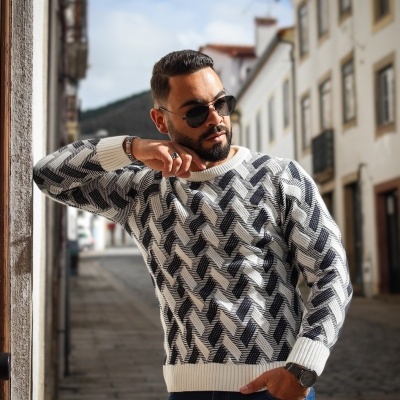 Homem de óculos escuros a usar camisola de malha cinza com padrão geométrico e calças azuis numa rua