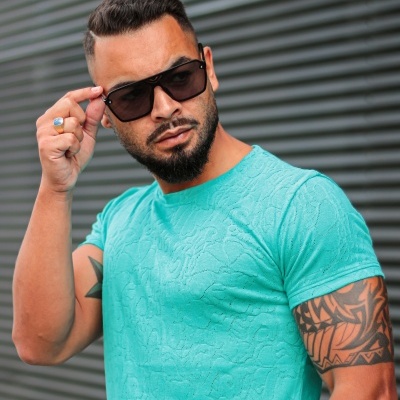 Homem com óculos de sol quadrados pretos e t-shirt azul clara com padrão texturizado em parede ripada cinza