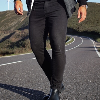 Homem a caminhar com calças pretas, botas e casaco cinzento numa estrada asfaltada com colinas ao fundo