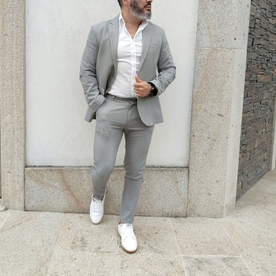 Homem com fato cinzento claro e sapatos brancos numa calçada em frente a parede branca
