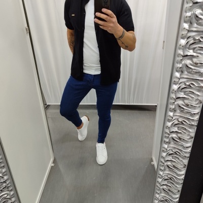 Homem vestido com camisa preta, camisola branca, calças azuis e sapatilhas brancas em frente a espelho grande com moldura prateada