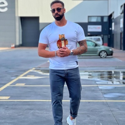 Homem de t-shirt branca com estampado de urso, calças azuis e sapatilhas brancas numa área industrial.