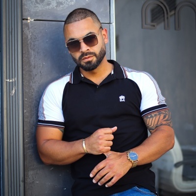 Homem usando polo preta e branca com logotipo e jeans azul encostado numa superfície metálica