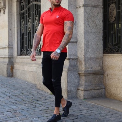 Homem com camisola polo vermelha, calças pretas e sapatos pretos numa rua com edifício de pedra ao fundo