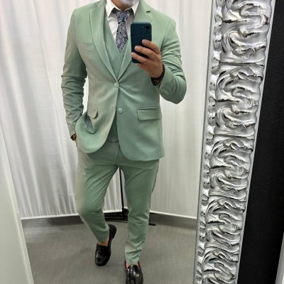 Homem com fato verde claro, camisa branca, gravata azul e branco, sapatos pretos em frente a espelho prateado